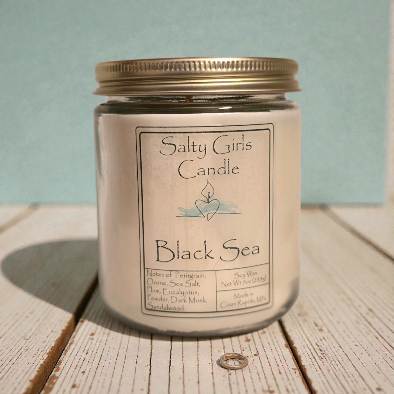 Candle jar labeled 'Salty Girls Candle Black Sea' on a wooden surface with a light blue background